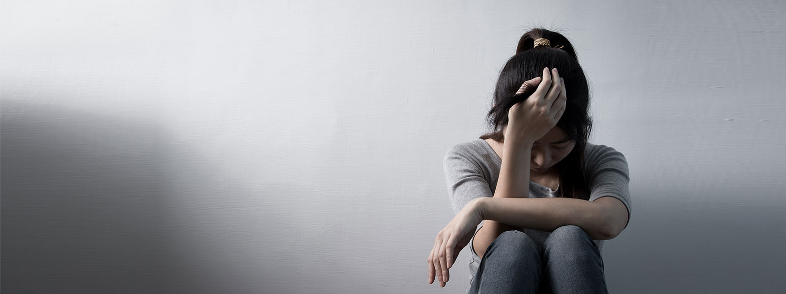 Young woman sitting on the floor against a white wall with her head resting on her hand, appearing sad or overwhelmed.