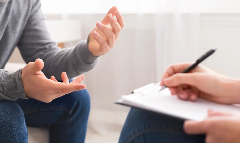 Close-up of a therapy session showing a person gesturing with their hands while another person takes notes on a clipboard.